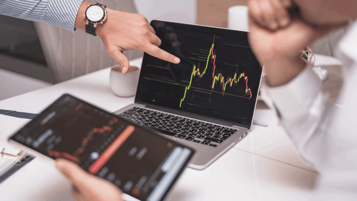 A laptop displaying a cryptocurrency stock chart with green and red candlesticks is on a white table. A hand with a watch is pointing at the screen. A tablet displaying a similar cryptocurrency chart is held in someone's hand.