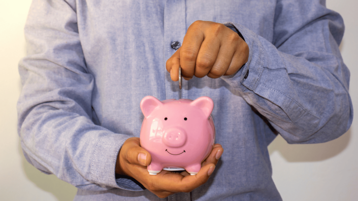 A person in a blue shirt places a coin into a pink piggy bank, symbolizing retirement planning and long-term savings.