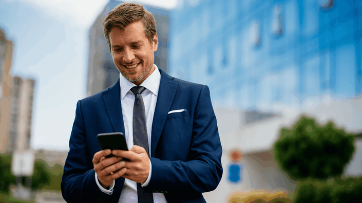 A smiling man in a blue suit and tie stands outdoors, looking down at his smartphone, presumably using a fintech banking app, with a modern glass building blurred in the background.