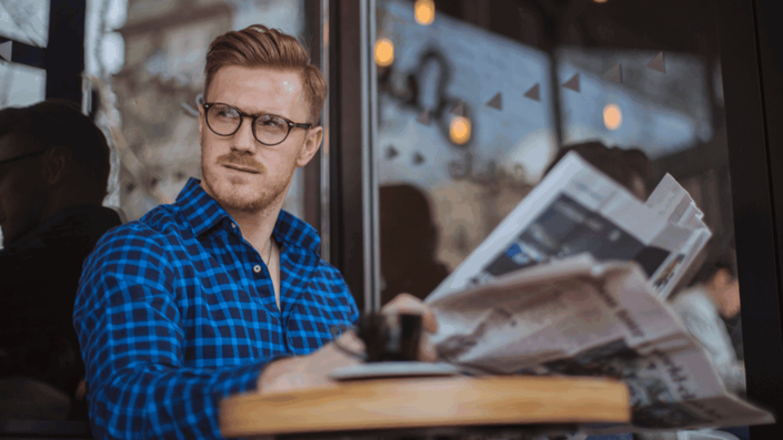 A man with red hair and a beard, wearing glasses and a blue plaid shirt, sits at a table outside a cafe, reading a newspaper about cryptocurrency and drinking coffee.