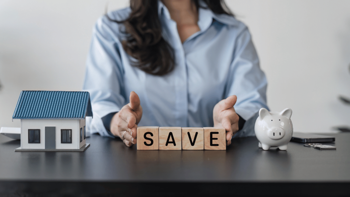 A person in a blue shirt sitting at a desk with a miniature house, a piggy bank, and wooden blocks spelling “SAVE,” representing financial planning and emergency funds management.
