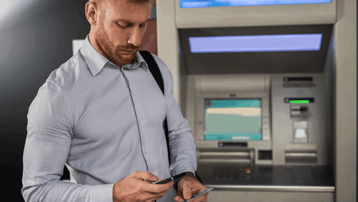 Man using mobile phone in front of an ATM, illustrating the transition from traditional banking to Cloud Banking.